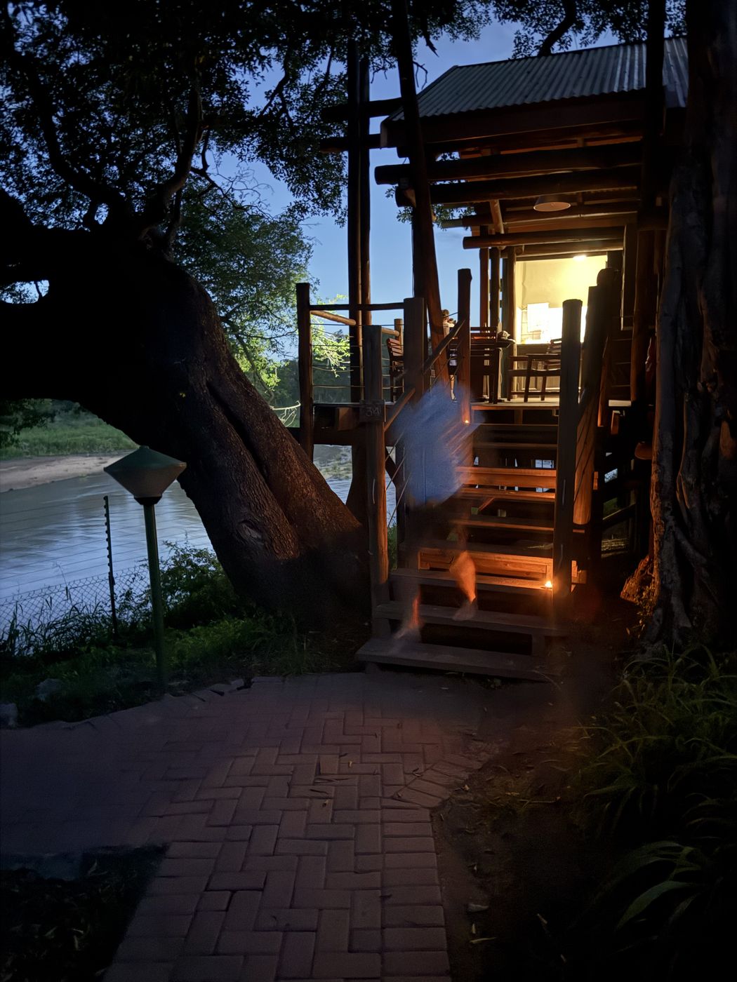 A SANParks camp unit lit from inside at dusk beside the river in South Africa